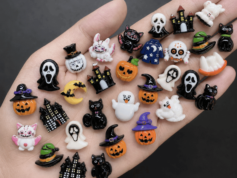 Cute Halloween resin charms with pumpkins, ghosts, black cats, haunted houses and witch hats displayed in a hand.