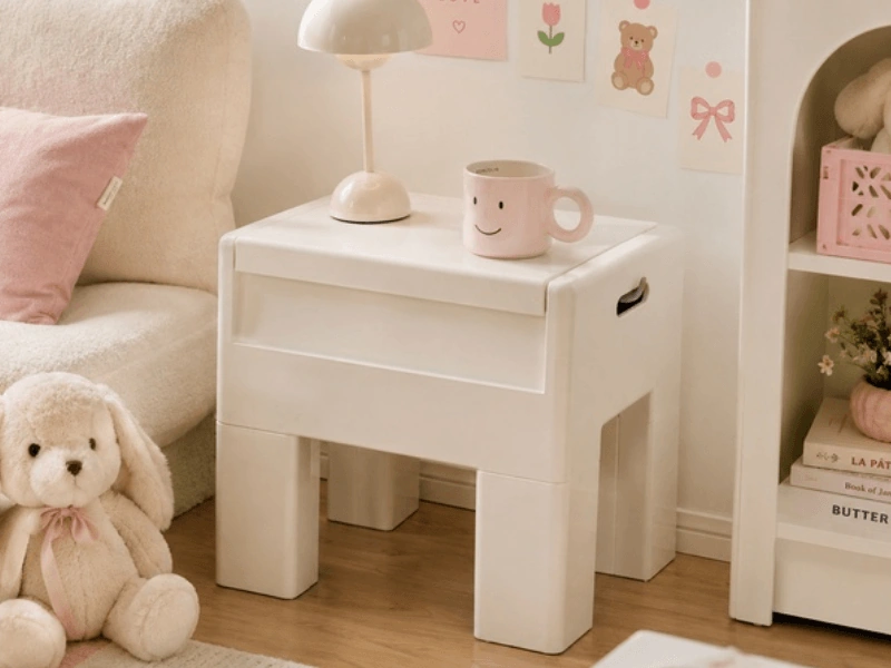 White plastic nightstand with a small lamp and pink smiley mug beside a cream chair in a cozy pastel bedroom.
