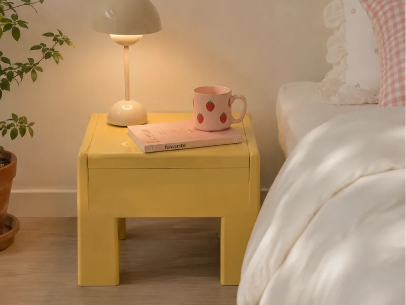 Yellow plastic nightstand beside a bed with a lamp, strawberry mug and book on top.