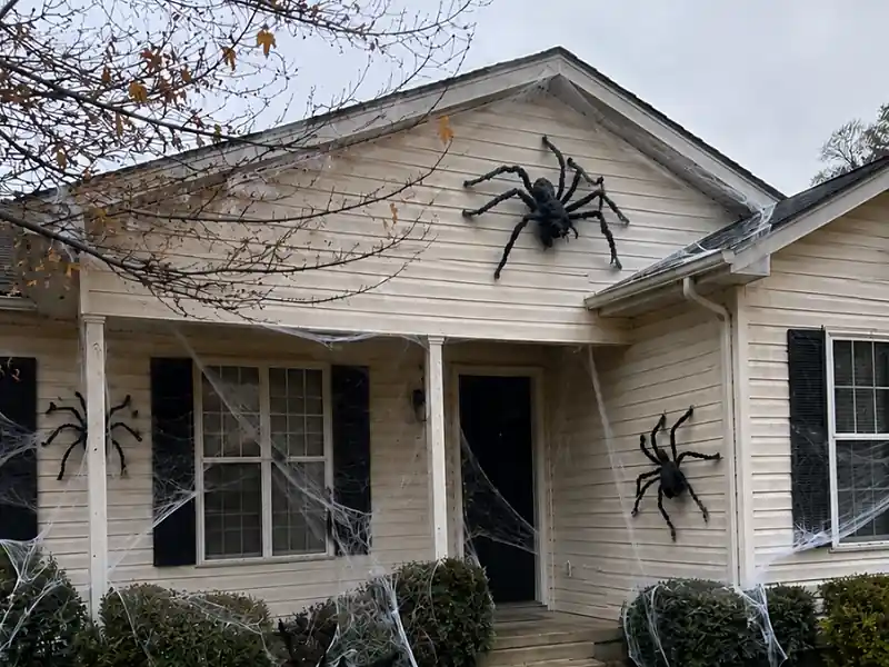 Giant black spider and fake spiderwebs covering a house for spooky outdoor Halloween party decorations.