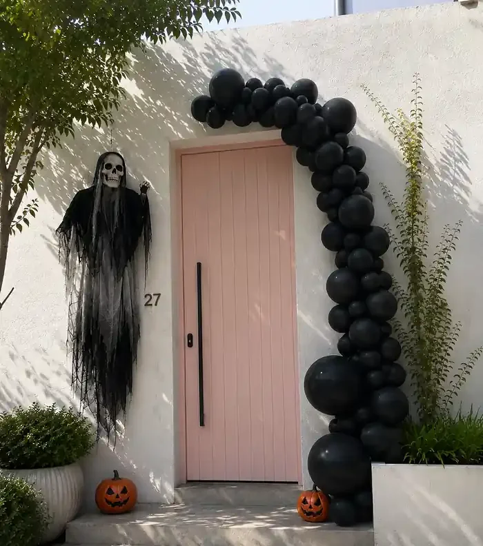 Hanging Grim Reaper beside a front door with black balloons and pumpkins for spooky outdoor Halloween party decorations.